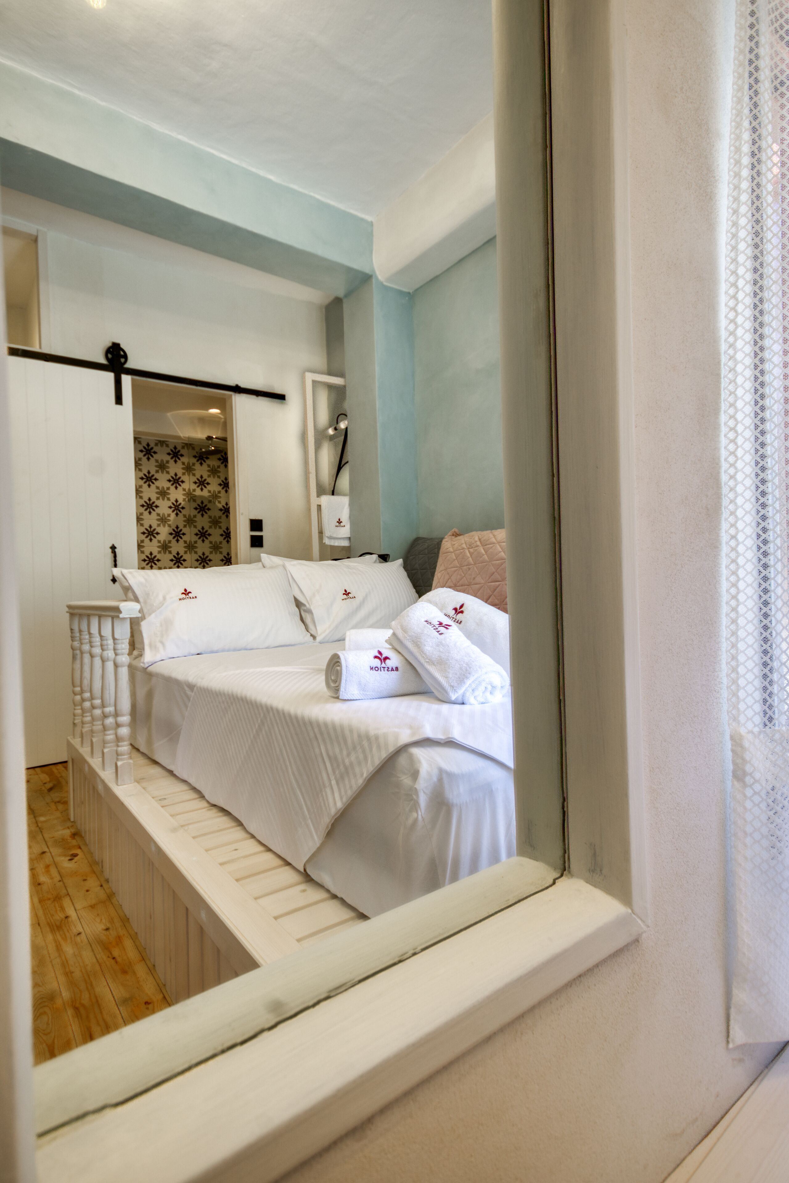 Een slaapkamer met een bed, een raam met gordijn, een houten vloer en een witte muur met lichtschakelaar.