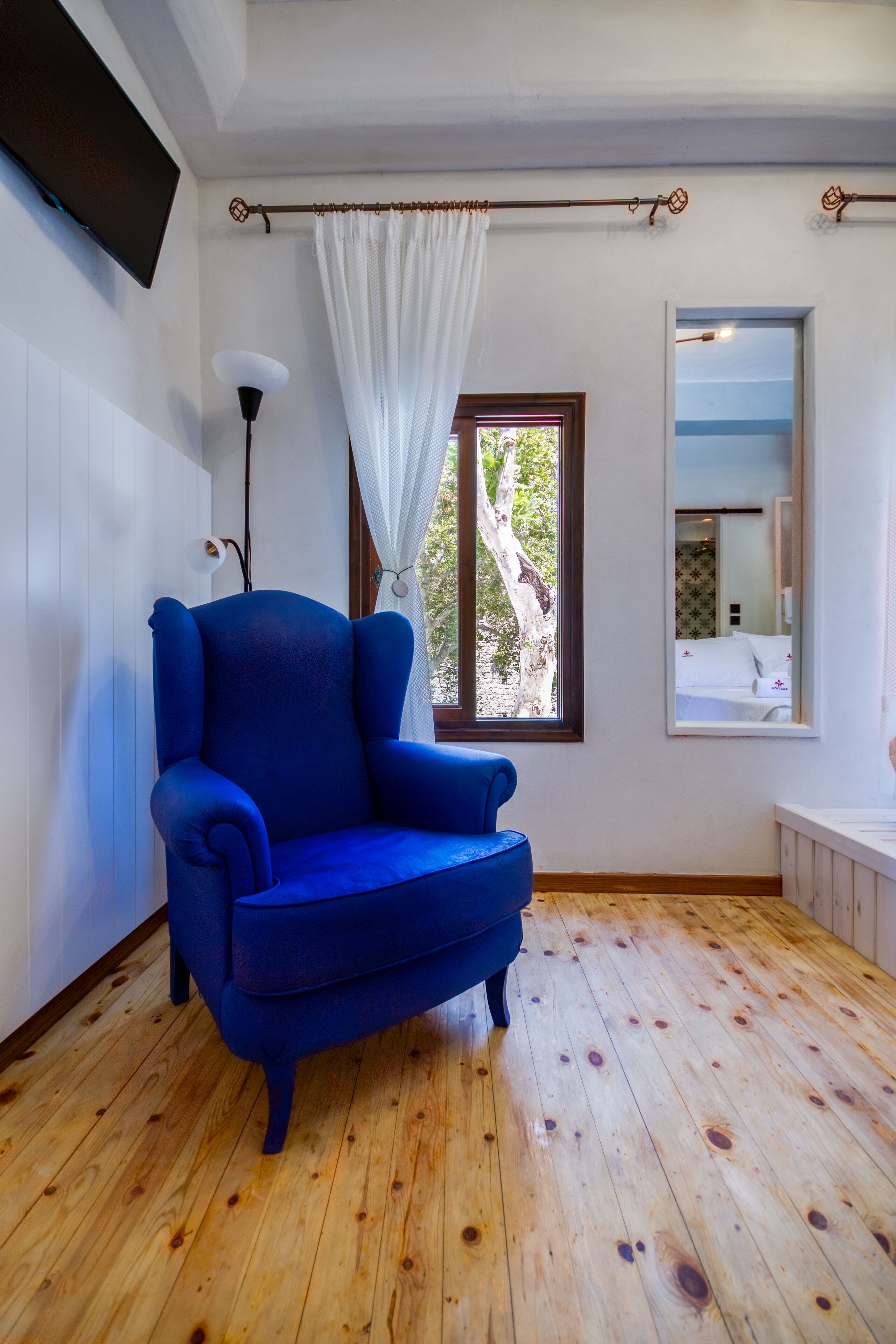 Een kamer met een blauwe fauteuil, houten vloeren, een raam met uitzicht op bomen, een wit gordijn en een spiegel aan de muur.