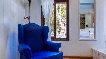Ein Zimmer mit blauem Sessel, Holzboden, einem Fenster mit Blick auf Bäume, einem weißen Vorhang und einem Spiegel an der Wand.