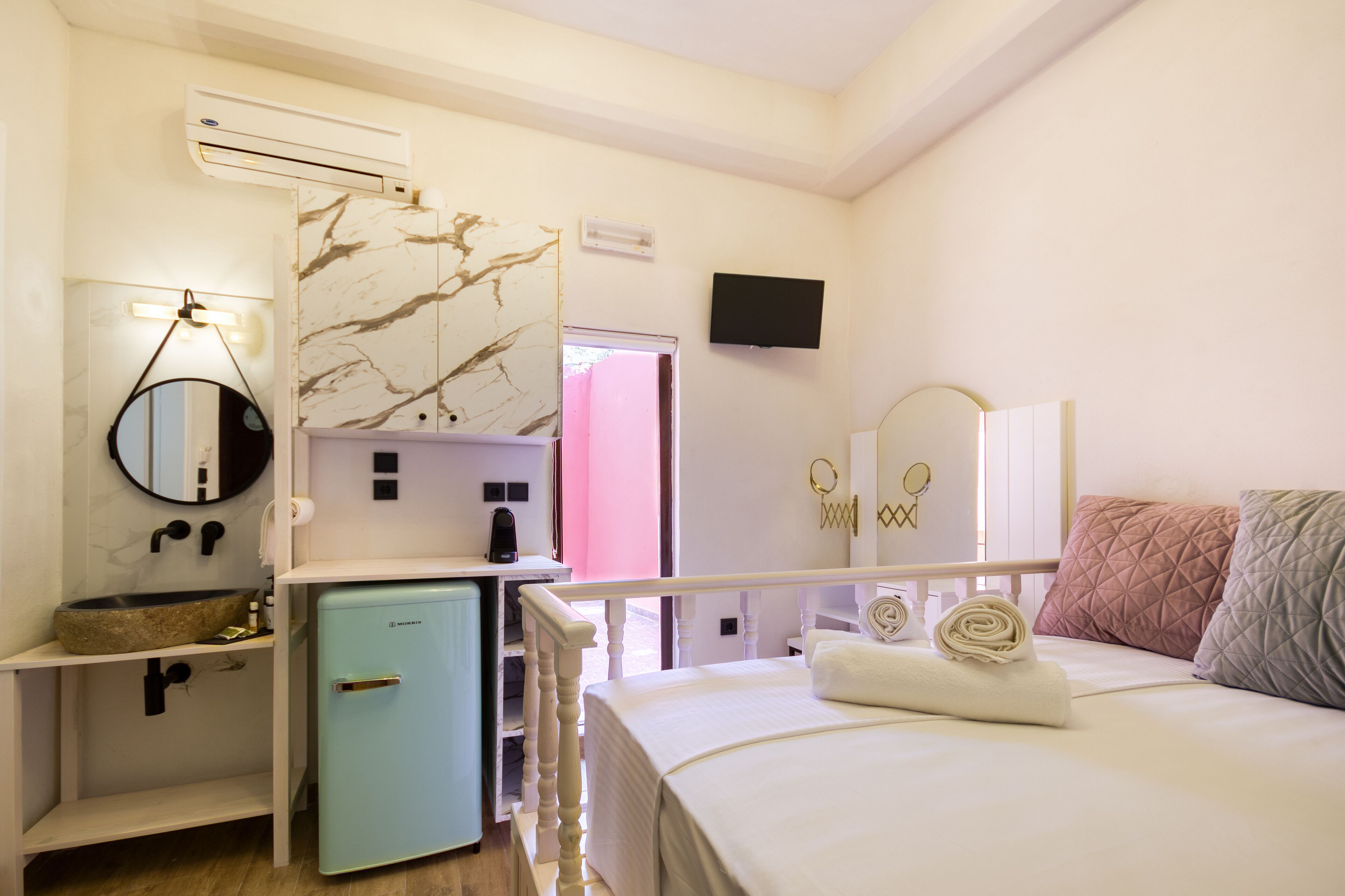 A hotel room with a bed, a small refrigerator, a sink, a mirror, a television, and a door leading to another room.
