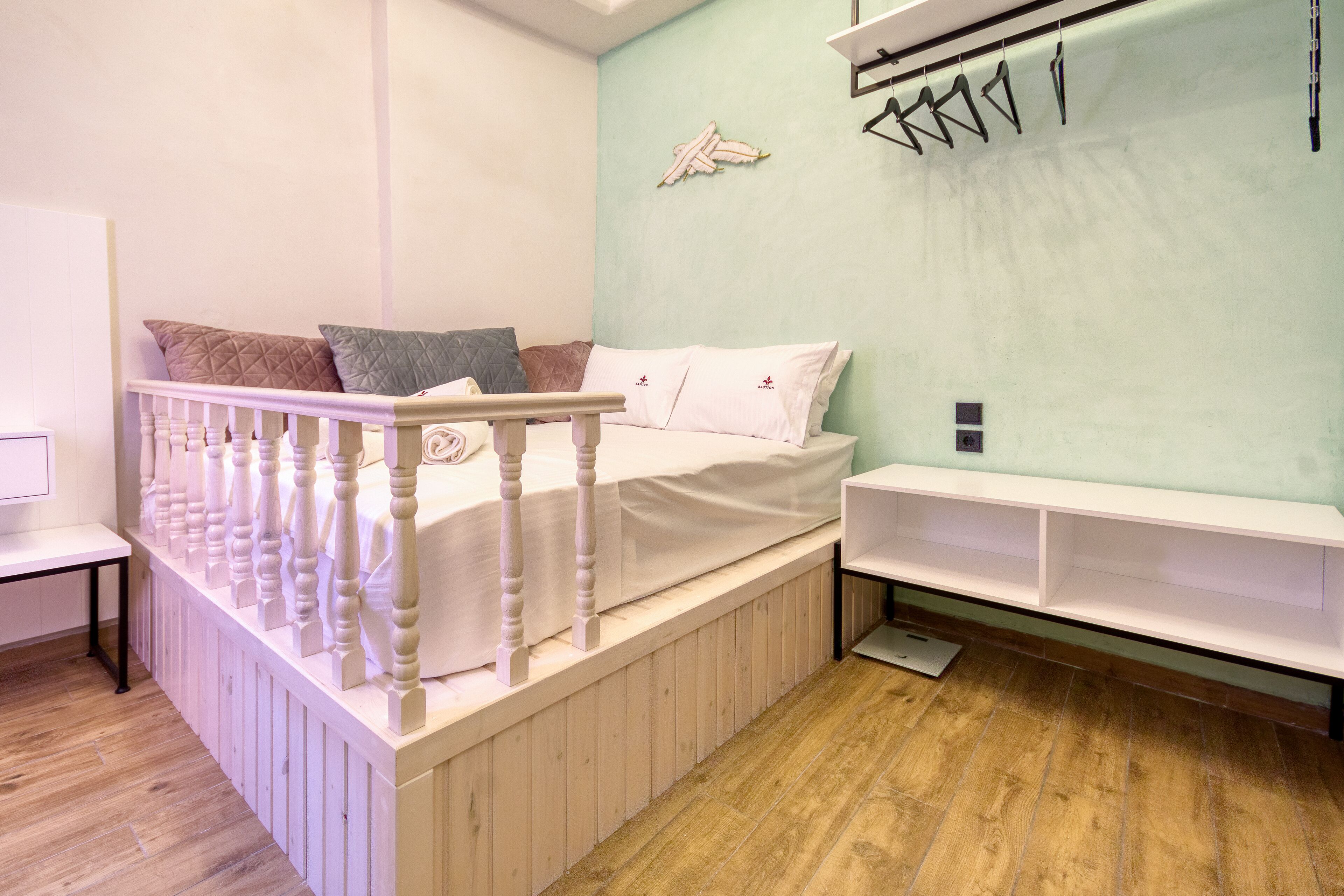 Un dormitorio con estructura de cama de madera, cabecera blanca y un banco empotrado. Hay una estantería contra la pared y una mesita de noche con una lámpara.