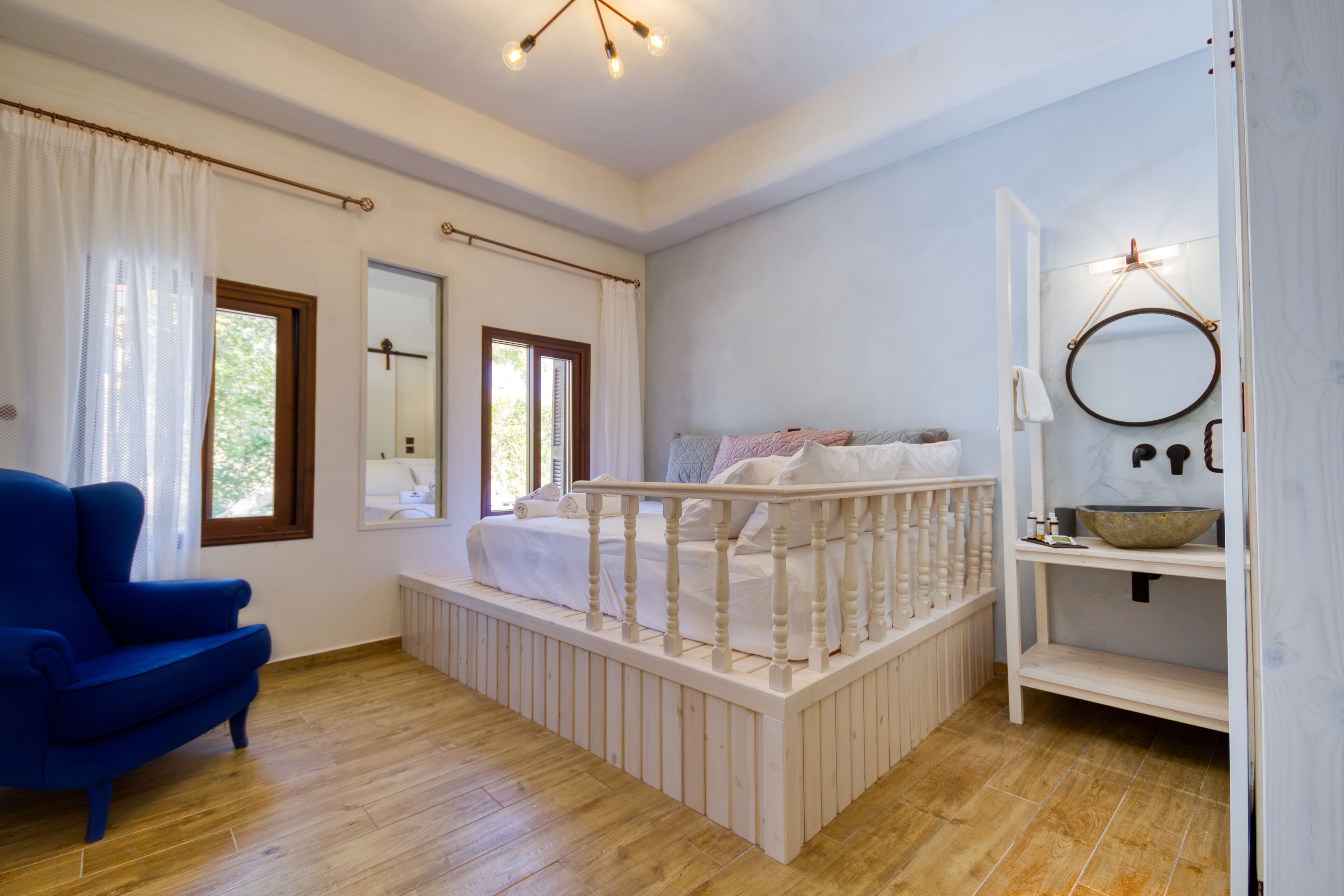 Un dormitorio con piso de madera, una silla azul, una cama con cabecera blanca, una cuna blanca, una ventana con cortinas y un baño con lavamanos y espejo.