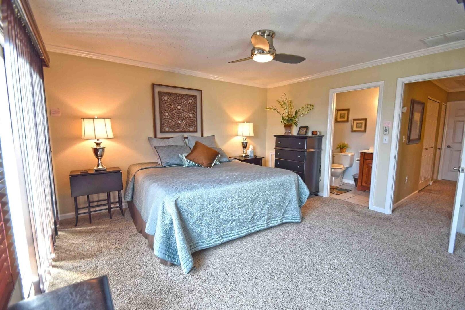 Une chambre à coucher comprenant un lit, deux tables de chevet avec des lampes, une commode, un ventilateur de plafond et une porte donnant sur une autre pièce.
