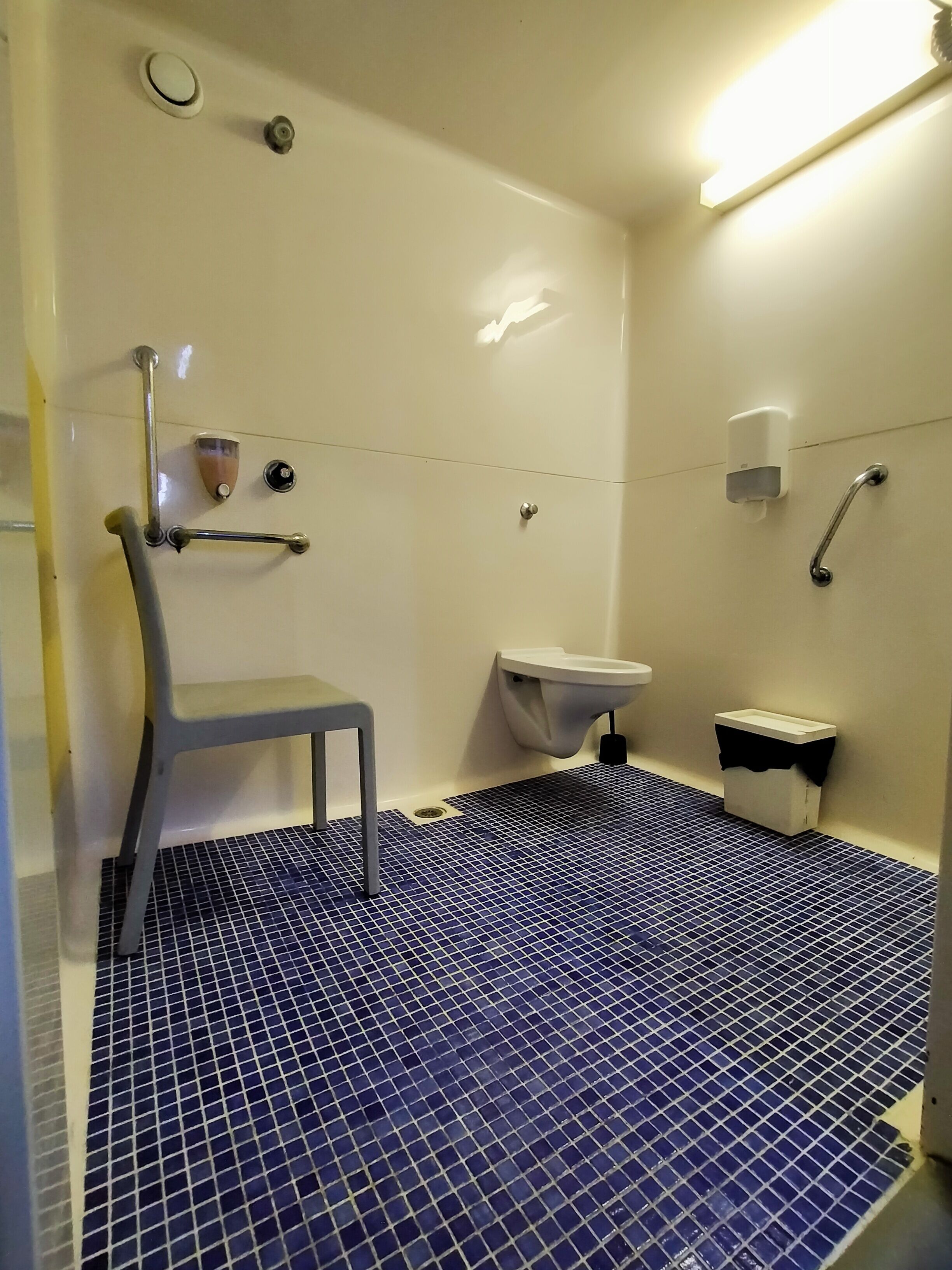 Un baño con piso de azulejos azules, un lavamanos blanco, un inodoro, una ducha con barra de apoyo y una silla.