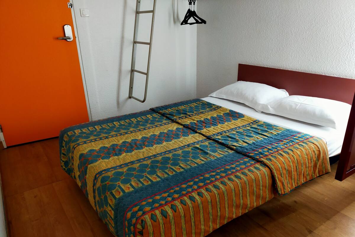 Un lit recouvert d’une couvre-lit à motifs, un plancher en bois, une porte orange et un mur blanc orné d’un porte-manteau mural.