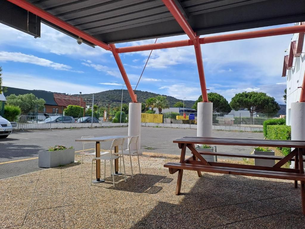 Un espace extérieur aménagé avec des tables et des chaises, une structure couverte, et une vue sur un parking et des bâtiments en arrière-plan.