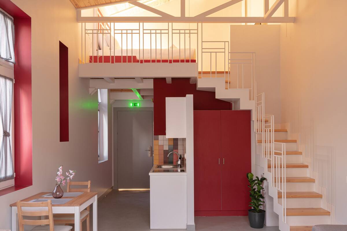 Un interior moderno con techo de madera, una escalera, una zona de comedor con mesa y sillas, y una cocina con armarios y pileta.