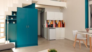 Un salón moderno con techo alto, un armario azul turquesa, una mesa de centro blanca y una zona de cocina con azulejos coloridos.