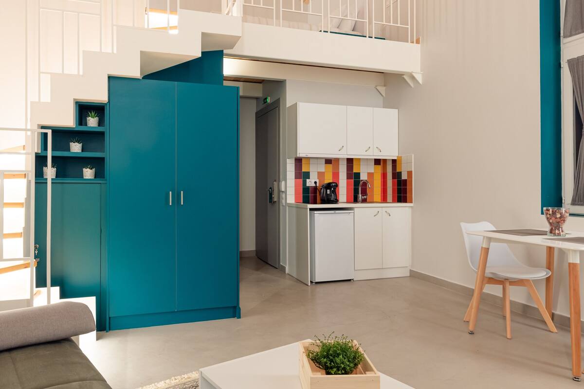 Un salón moderno con techo alto, un armario azul turquesa, una mesa de centro blanca y una zona de cocina con azulejos coloridos.