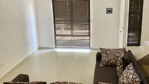 A living room with a sofa set, a coffee table, and a patterned throw pillow. There is a window with blinds, a door, and a light switch on the wall.