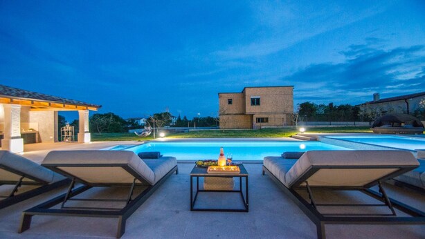 A pool area with lounge chairs and a table, a building in the background, and a clear sky. - Villa Mabuhay (Marcana)
