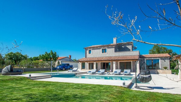 A two-story house with a swimming pool, outdoor seating, and a blue car parked nearby.