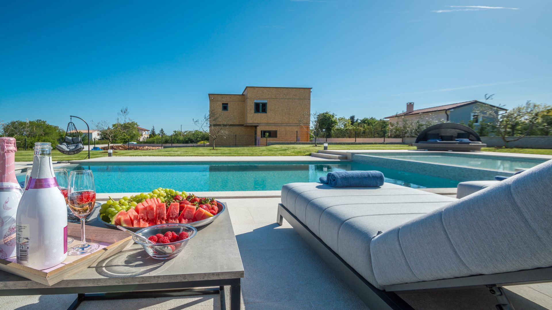Un espace aménagé au bord de la piscine, avec un fauteuil de relaxation, une table sur laquelle se trouve un panier de fruits et une bouteille d’eau.