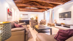 A modern living room with a high ceiling, wooden beams, a flat-screen TV, and a dining area in the background. - Villa Lucia (Marcana)