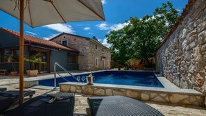 Una piscina con sillas de descanso y una sombrilla, rodeada de edificios de piedra y árboles.