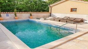 Una piscina rectangular con agua azul cristalina, rodeada por un solárium con baldosas. Se observan sillas de descanso y un pequeño edificio con ventanal al fondo.