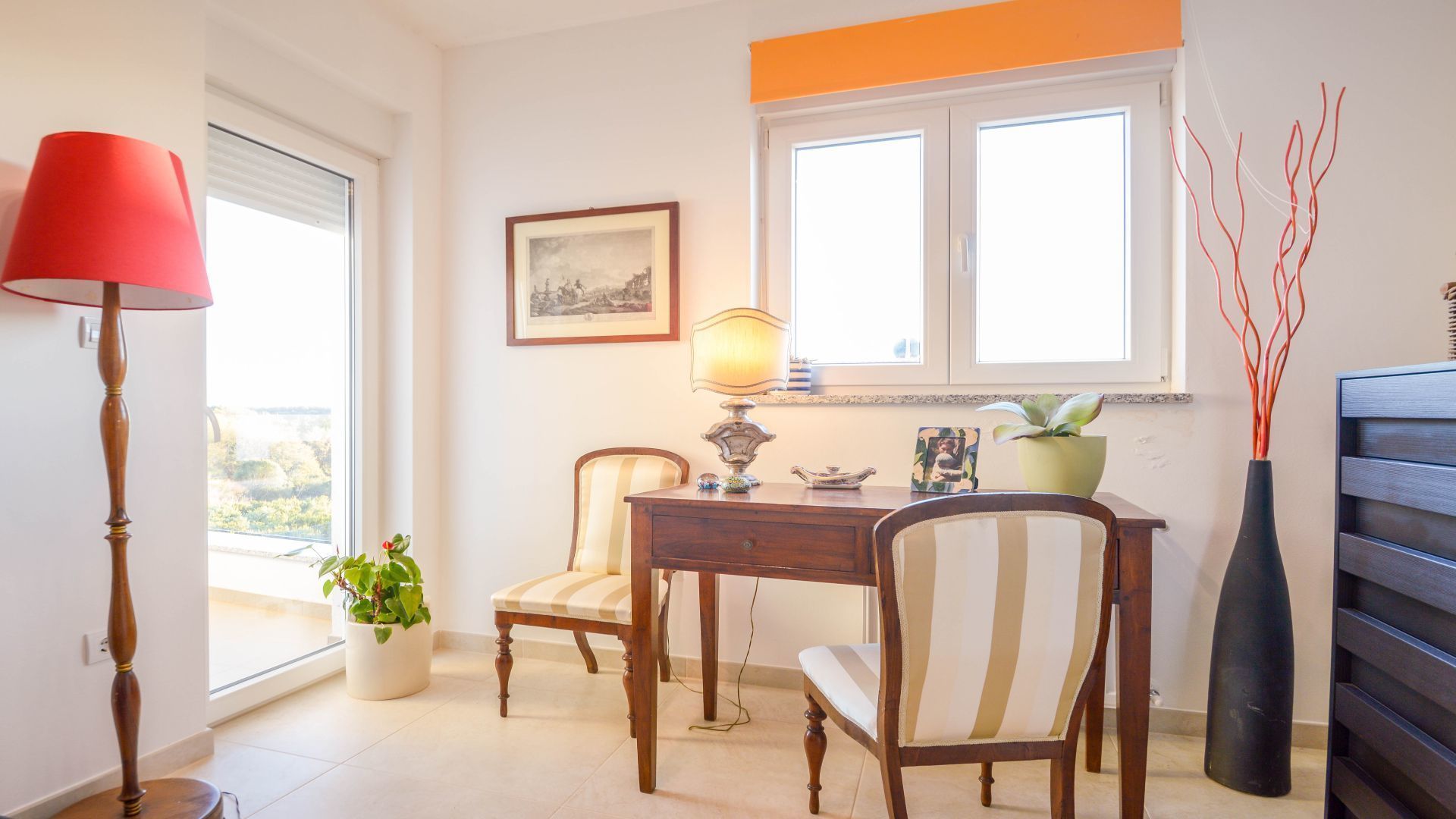 A room with a wooden desk, two chairs, a lamp, a potted plant, and a framed picture on the wall.