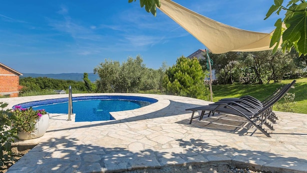 A swimming pool with a stone deck, surrounded by greenery and a clear sky.
