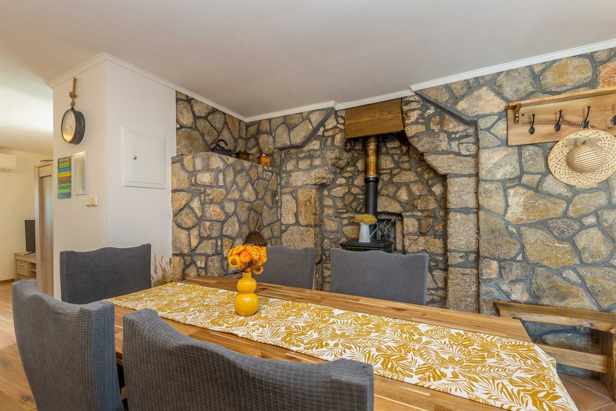 Un coin repas avec un mur en pierre, une table en bois agrémentée d’un chemin de table à motifs et des chaises à revêtement gris.