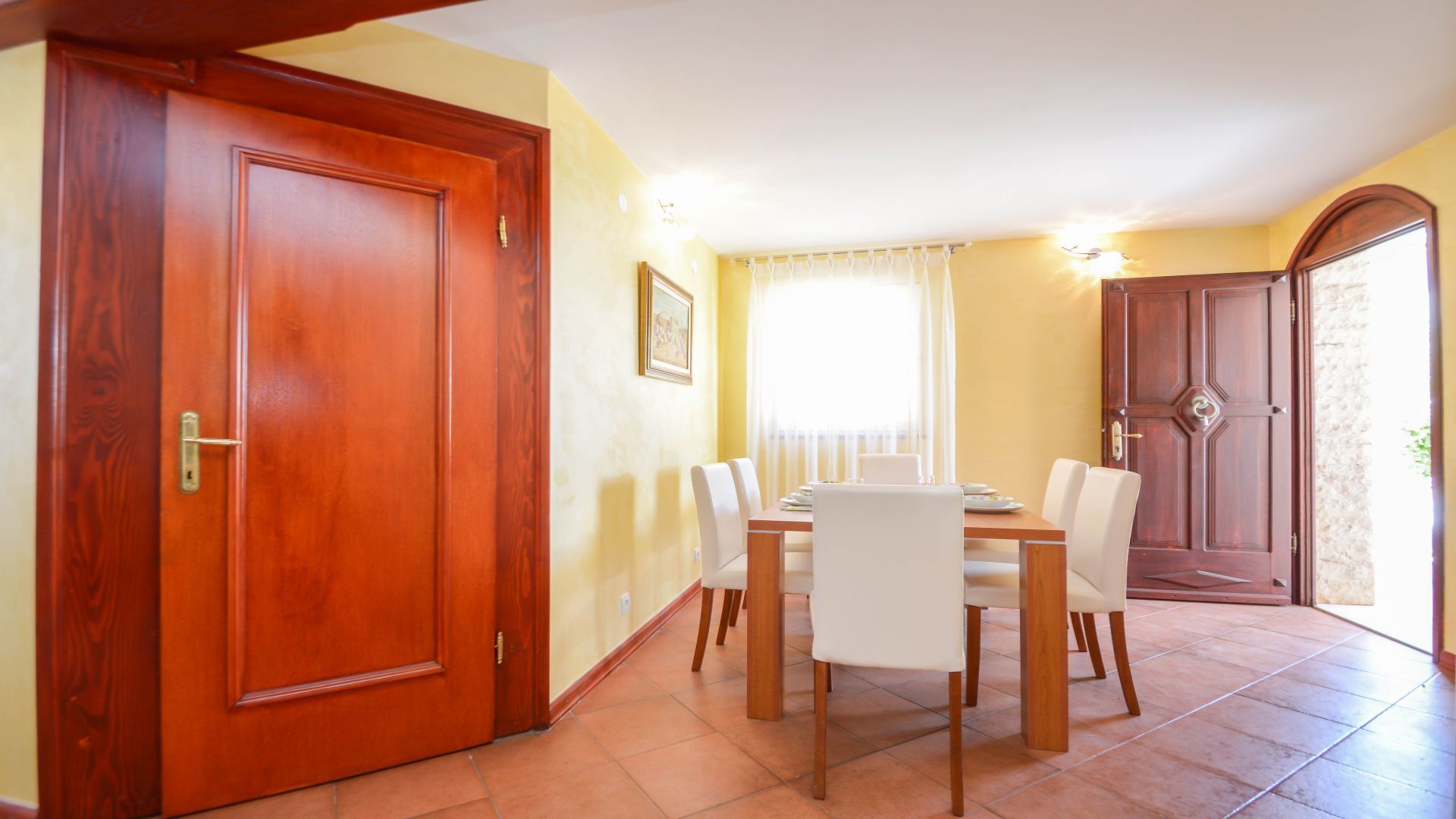 A dining area with a wooden table and chairs, a window with curtains, and a door with a brass handle.