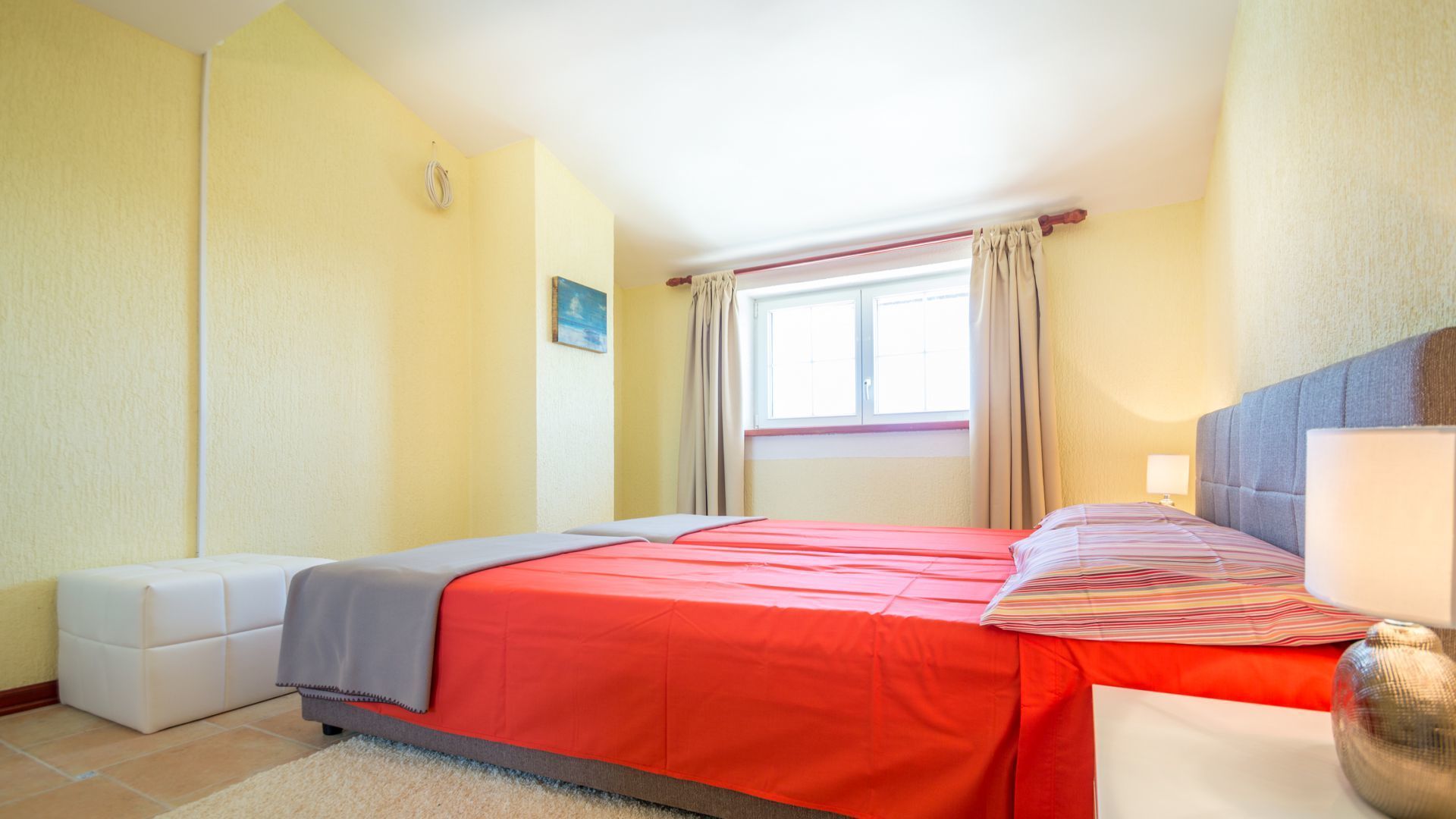 A hotel room with a bed, a bedside table, a lamp, a window with curtains, and a small painting on the wall.
