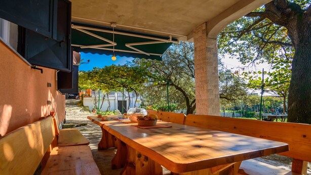 An outdoor patio with wooden benches and a table, surrounded by trees and a clear sky. - Holiday House Roses (Medulin)