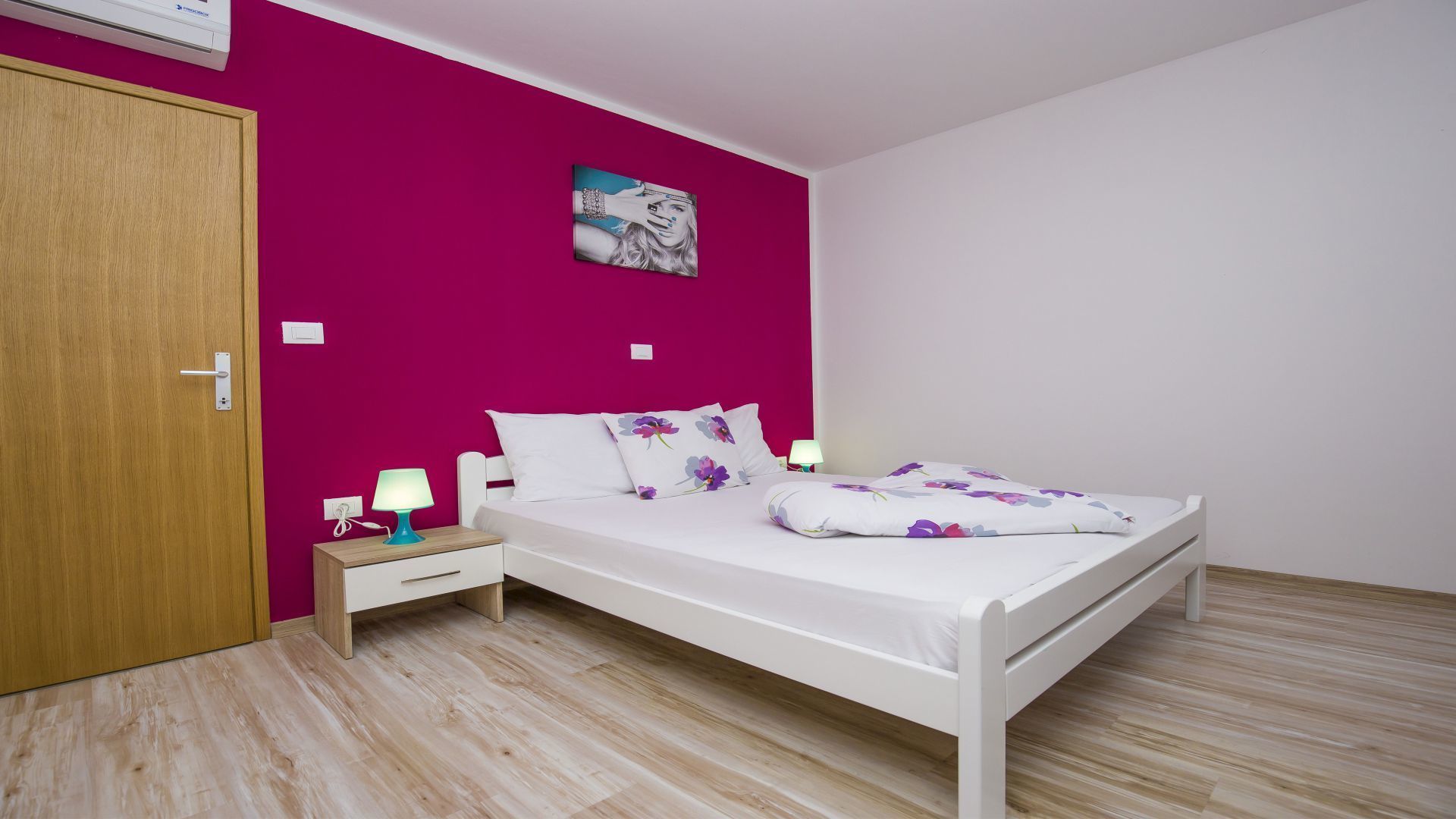 Un dormitorio con una cama blanca, una puerta de madera, una mesita de noche con una lámpara y una pared decorada con un cuadro.