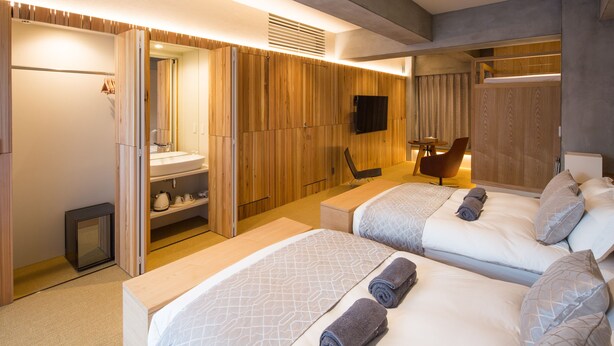 A modern hotel room with two beds, a sitting area, a bathroom with a mirror and sink, and a television mounted on the wall. - Sakura Cross Hotel Tokyo Kayabacho (Tokyo)