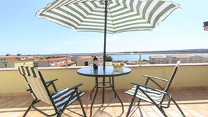 Una terraza con una mesa redonda, dos sillas a rayas, un paraguas a rayas y vistas al mar y edificios en el fondo.