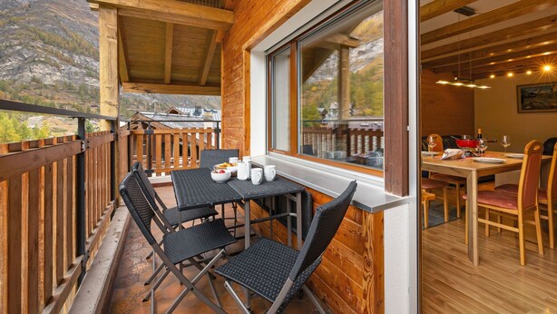 A wooden deck with a table and chairs, a window with a view of mountains, and a dining area with a table set for two. - Silence by Interhome (Zermatt)