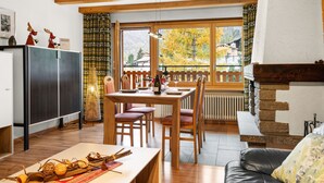 A cozy living room with a wooden table, chairs, a fireplace, and a view of autumn trees. - Silence by Interhome (Zermatt)