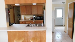 A kitchen with wooden cabinets, a stainless steel sink, a microwave, and a refrigerator. There are two stools at the counter.