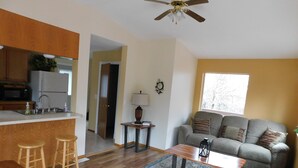 A modern living room with a sofa, coffee table, and wooden flooring. A kitchen area with a microwave and sink is visible in the background.