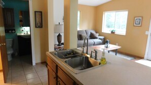 A kitchen with a sink, cabinets, and a window. There is a living room with a couch and a window.