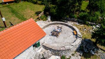 Un patio de piedra circular con un pozo para fuego y asientos, rodeado por un muro de piedra y un edificio con techo rojo.