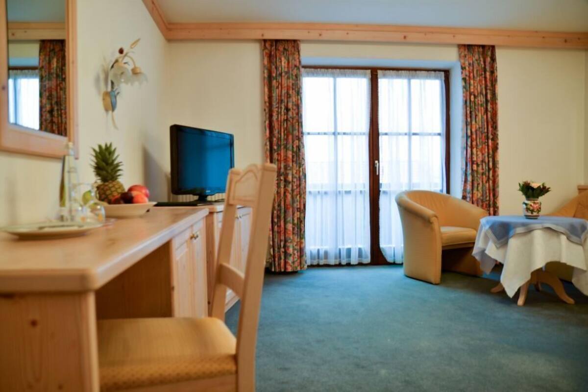 A room with a wooden desk, a television, a chair, a table with a vase, and a window with curtains.