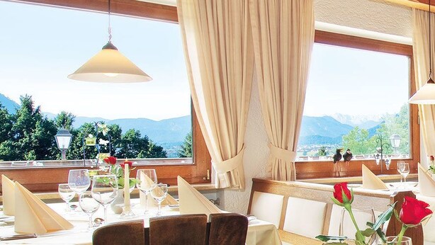 A restaurant with multiple tables, wooden beams, large windows with mountain views, and hanging pendant lights. - Hotel Restaurant Alpenhof (Uebersee)