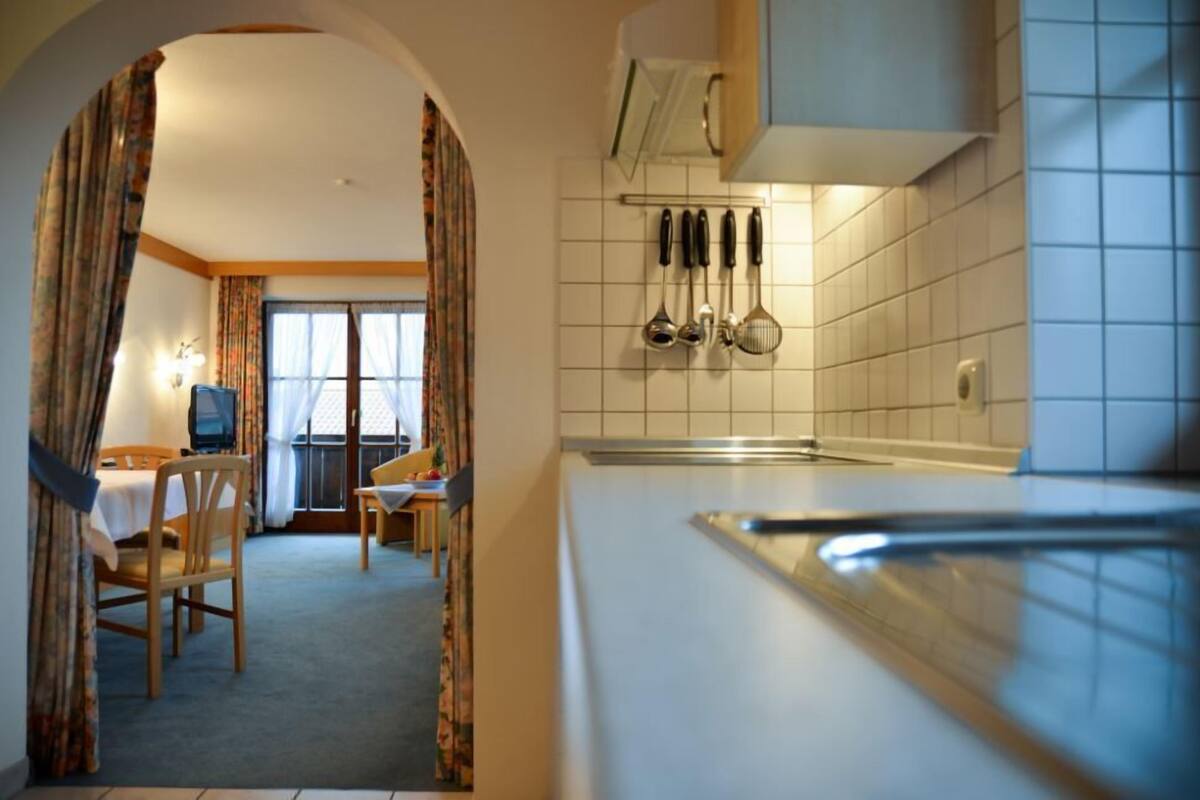 A room with a dining area, a kitchen with utensils hanging on the wall, and a window with curtains.
