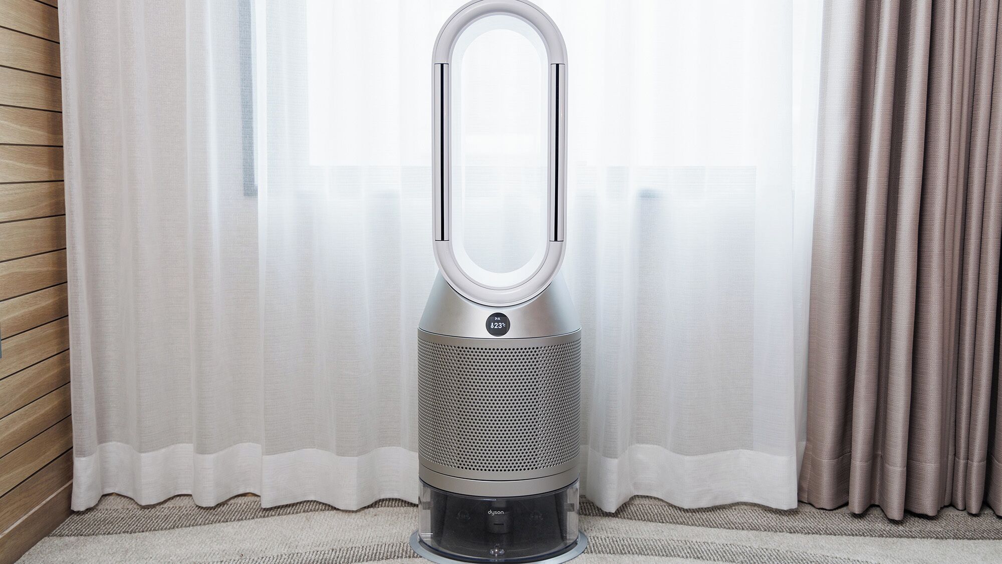 A Dyson bladeless fan in a room with a wooden floor and a window with sheer curtains.