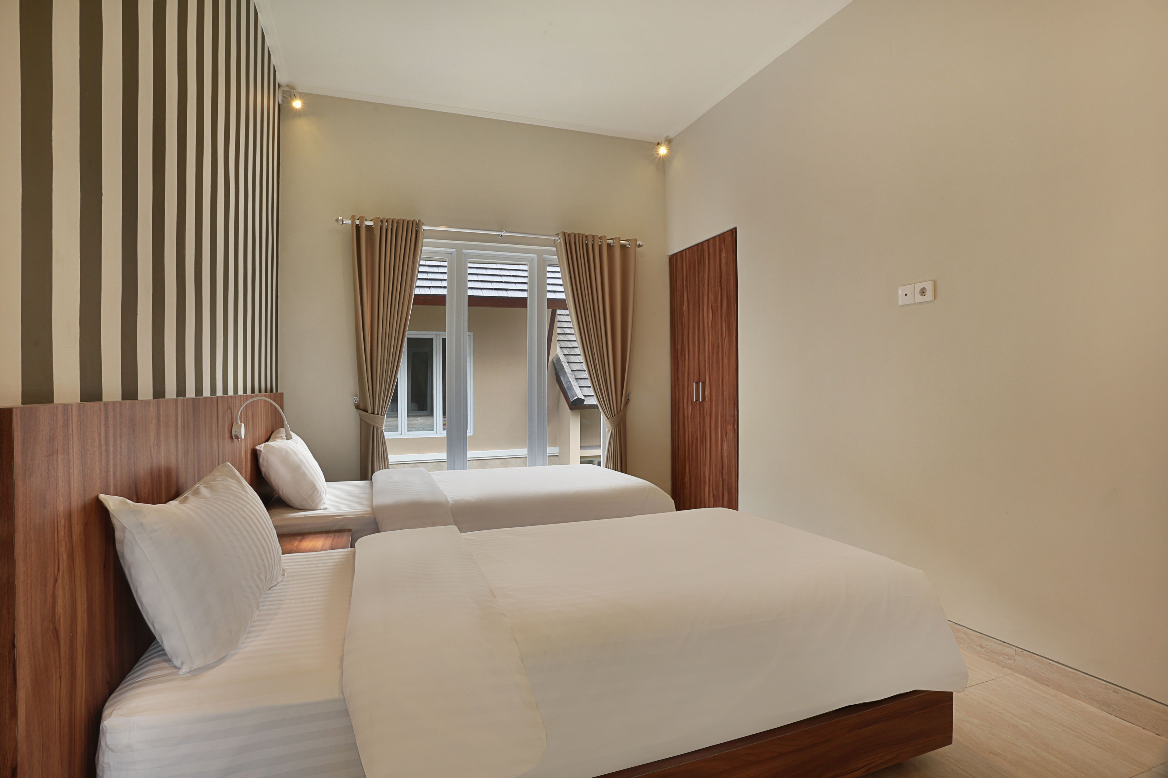 A hotel room with two beds, a wooden headboard, a window with curtains, and a view of a staircase.