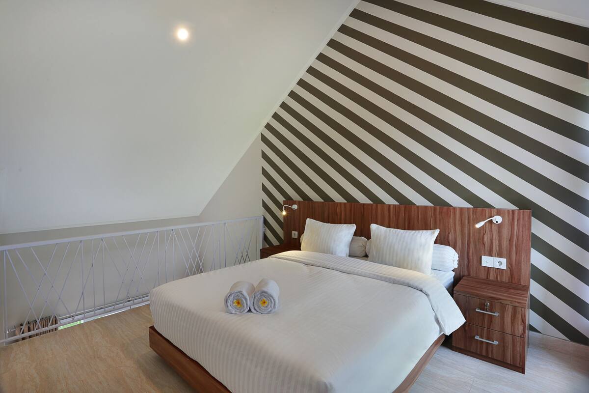 Een slaapkamer met een schuine kap, een bed met netjes gevouwen handdoeken, een houten nachtkastje en een gestreepte wand.