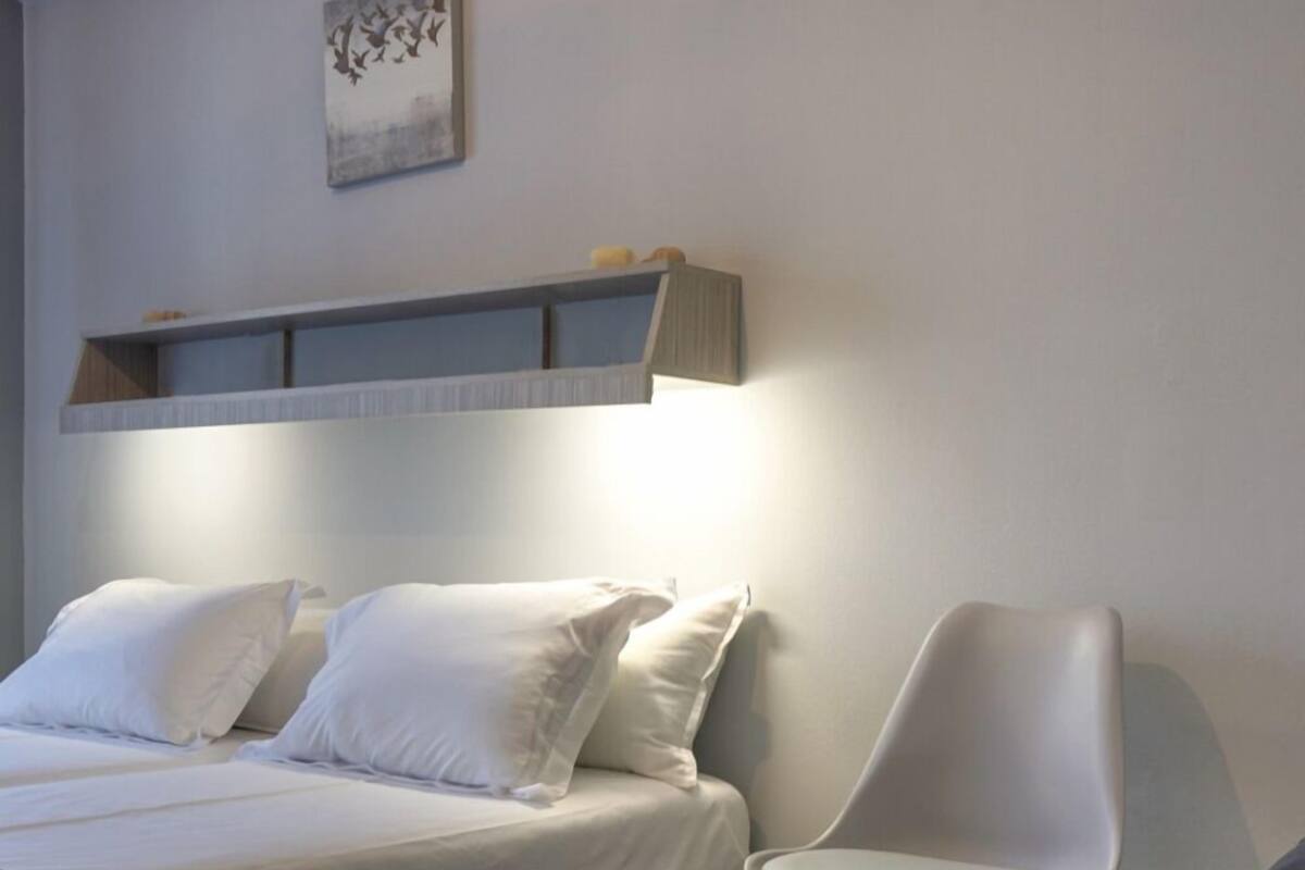 Un dormitorio moderno con una cama de madera, ropa de cama blanca, una silla gris y un estante montado en la pared.