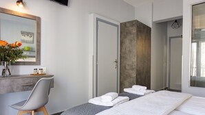 A modern hotel room with a bed, a desk, and a chair. There is a mirror on the wall, a vase with flowers, and a door leading to another room. - Liprando (Paros)