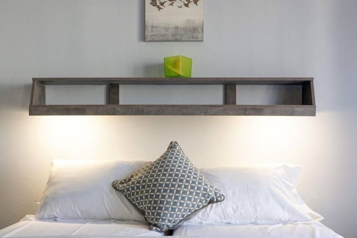 Una cama bien tendida con sábanas blancas y una almohada estampada, una manta texturizada gris y toallas dobladas en el suelo.