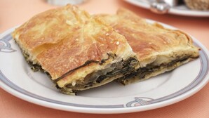 A plate with a piece of layered pastry, possibly spanakopita, on a table. - Liprando (Paros)