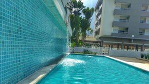 A swimming pool with clear blue water, surrounded by a tiled wall and a building with balconies. - Beautiful new apartment! (Ubatuba)