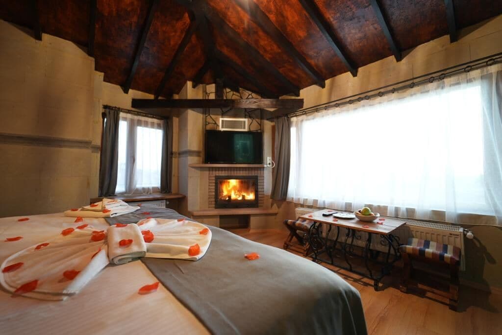A cozy room with a fireplace, a bed with rose petals, a table with a bowl of fruit, and a window with curtains.