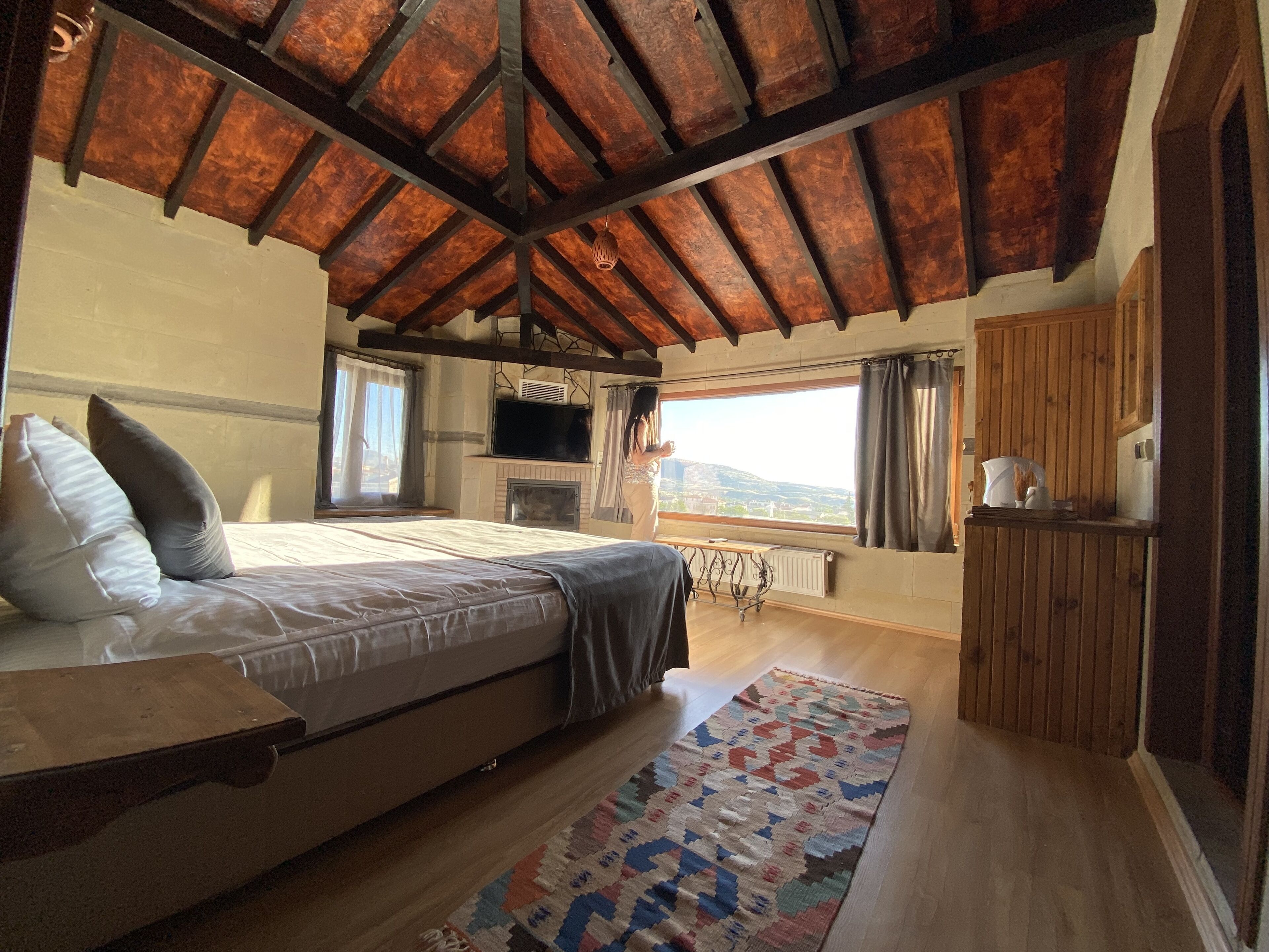 A bedroom with a wooden ceiling, a bed with pillows, a TV, a fireplace, and a view of the outdoors.