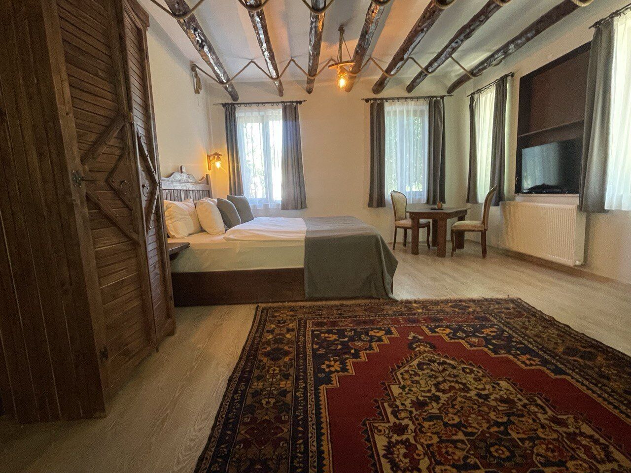 A room with a bed, a table, and a chair. There is a window with curtains and a radiator.
