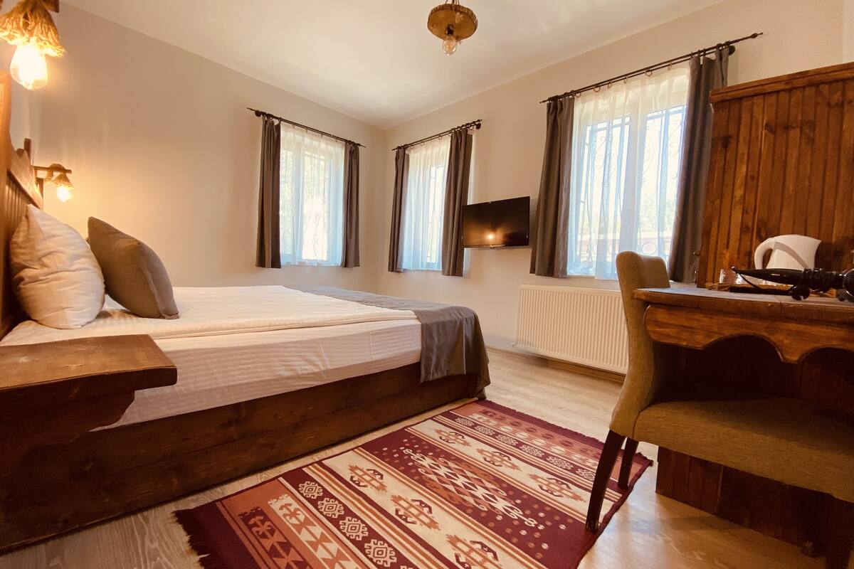 A bedroom with a bed, a desk, and a chair. There are windows with curtains, a radiator, and a television on the wall.